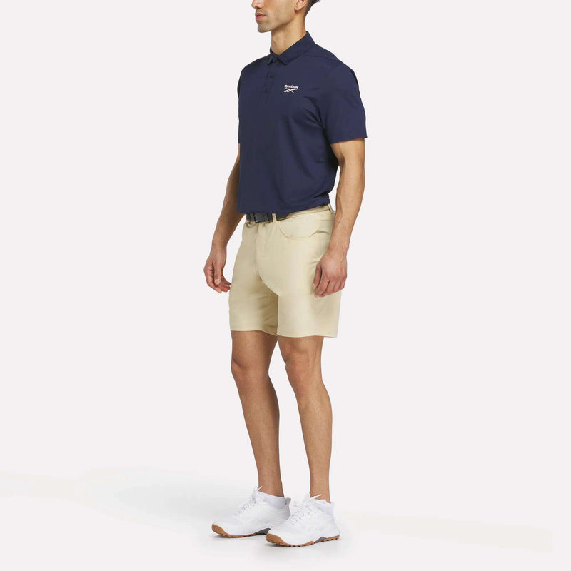 A man stands against a white background wearing a navy polo, Men's Favorite Golf Shorts 8.5" by Reebok in beige with RBK-DRY warp knit fabric, a black belt, and white sneakers.