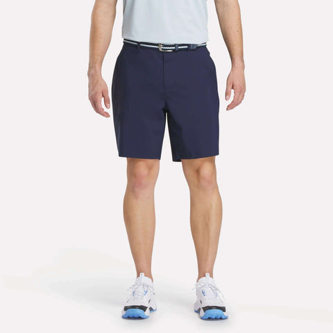 A man stands against a plain white background wearing a light blue shirt, Reebok Men's Favorite Golf Shorts 8.5" in dark blue, a black-and-white belt, and white athletic shoes with blue soles.