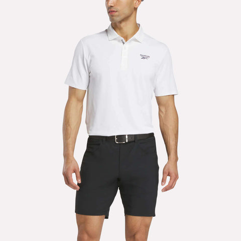 A man in a white short-sleeve polo with a chest logo, Reebok Men's Favorite Golf Shorts 8.5" in black, and a black belt stands against a plain light background.