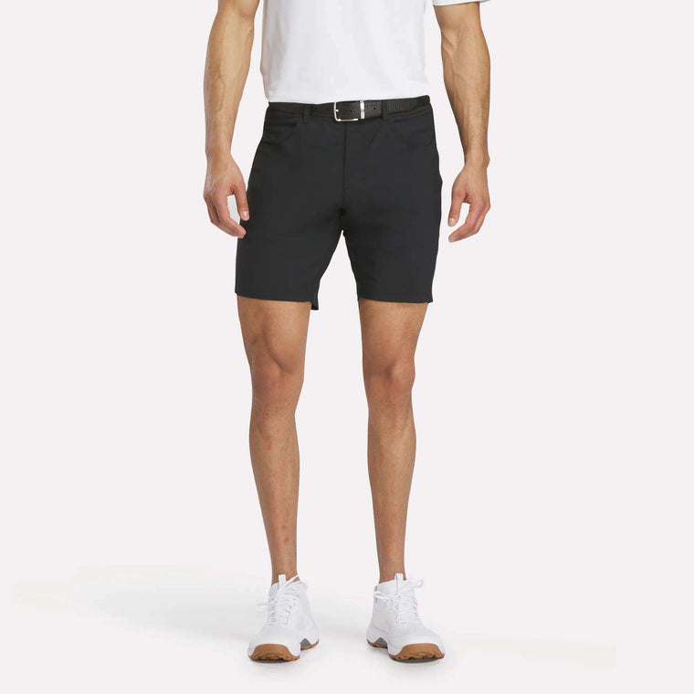 A man models Reebok Men's Favorite Golf Shorts 8.5", paired with a white polo, black belt, and white sneakers against a plain white background; only his body from the shoulders down is shown.