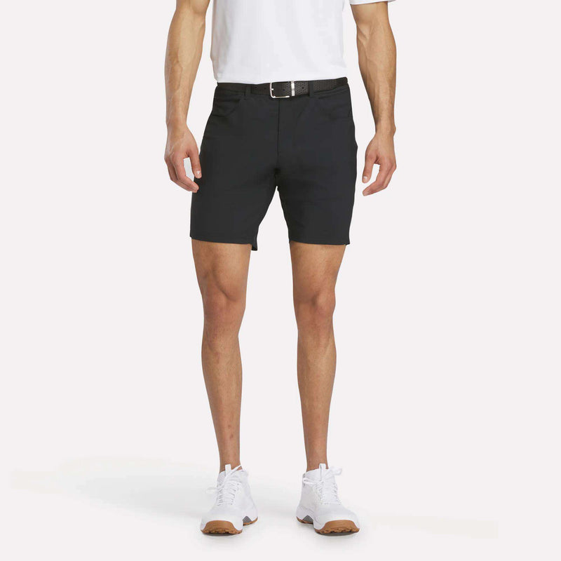 A person wears Reebok Men's Favorite Golf Shorts 8.5" made from moisture-wicking, stretchy nylon-spandex, along with a white polo, black belt, and white sneakers. The image is cropped at the shoulders against a plain white background.