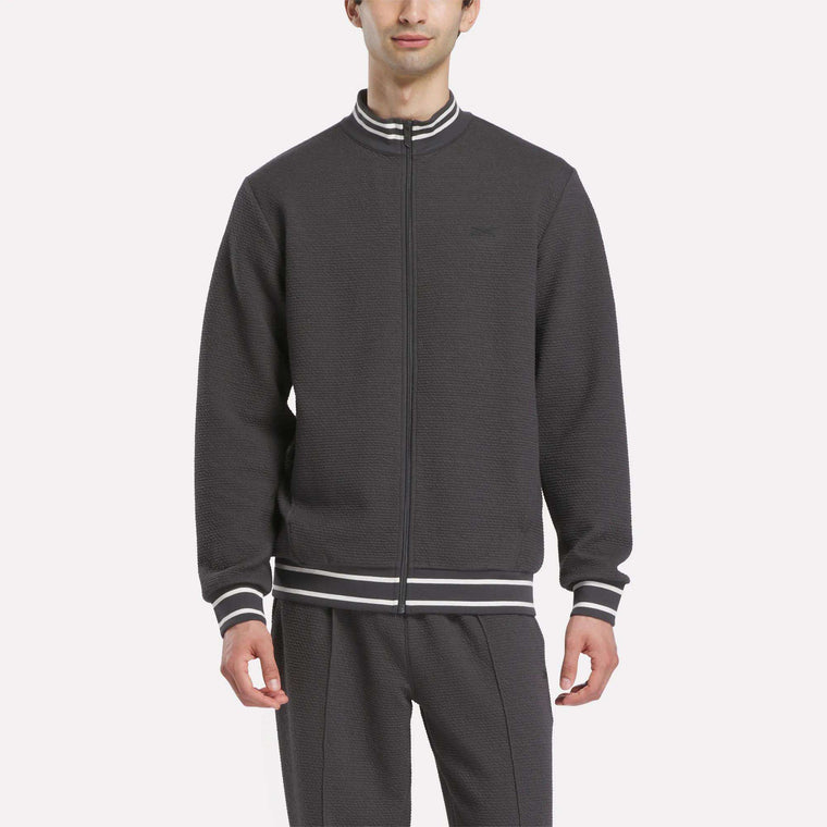 A man wears the Reebok Men's Club Sport Textured Jacket with matching pants, featuring white and gray striped cuffs and collar, standing against a plain light background.