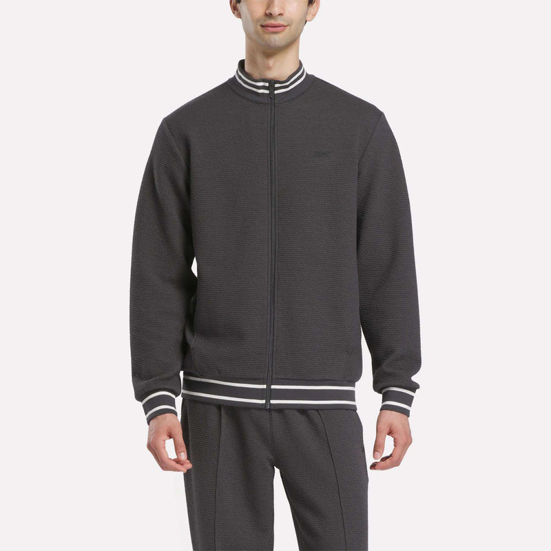 A man wears the Reebok Men's Club Sport Textured Jacket with matching pants, featuring white and gray striped cuffs and collar, standing against a plain light background.