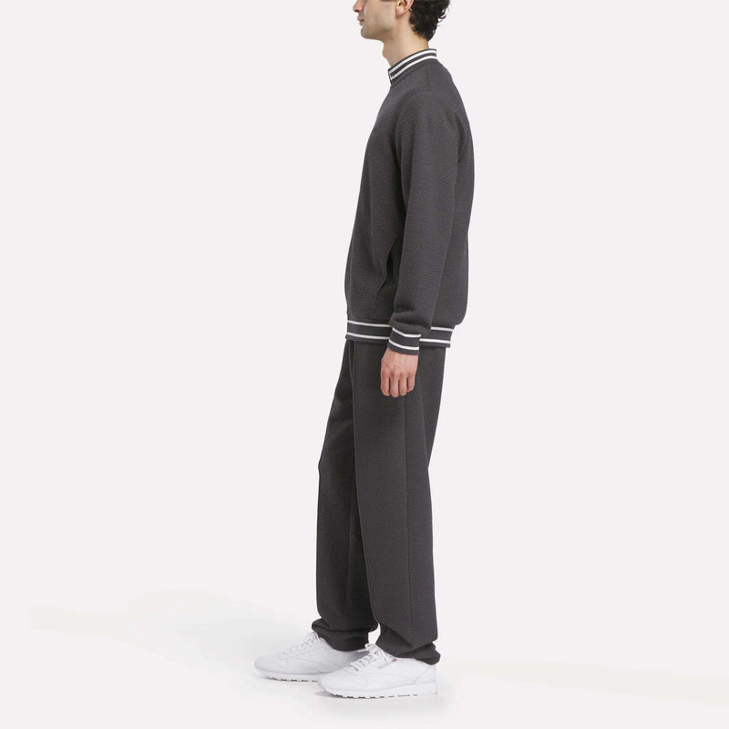 A man in profile wears the Reebok Men's Club Sport Textured Jacket in dark gray with white-striped cuffs and collar, paired with white sneakers, standing against a plain white background.