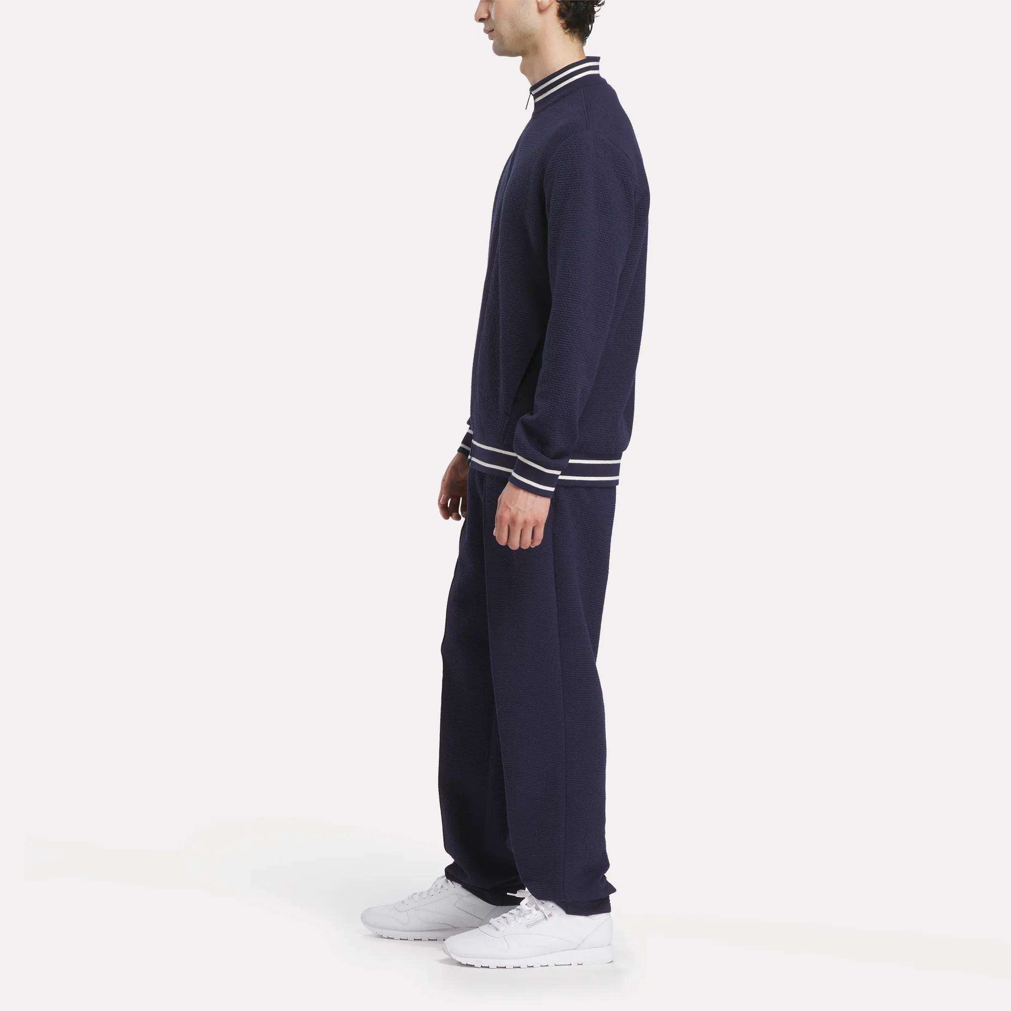 A man in profile wears a sporty navy tracksuit with white-striped cuffs and collar, featuring the Reebok Men's Club Sport Textured Jacket and white sneakers, standing against a plain white background.
