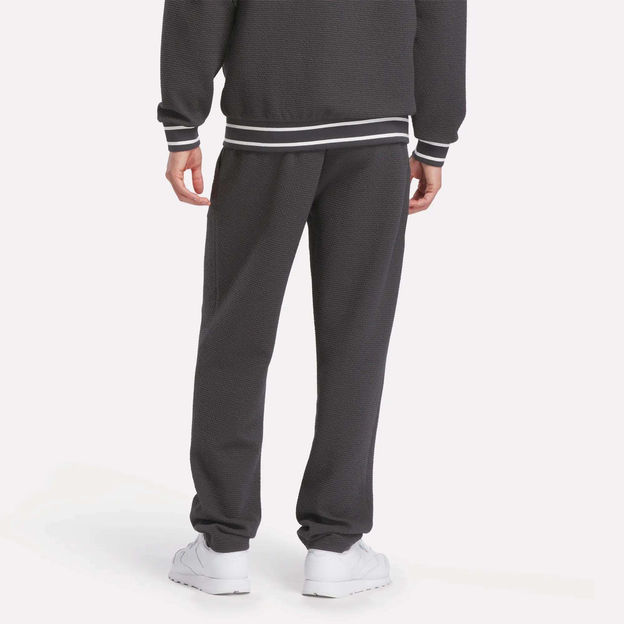 A person stands facing away, wearing Reebok Men's Club Sport Textured Pants with a slim tapered fit and white-striped cuffs, paired with white sneakers, set against a plain light background.