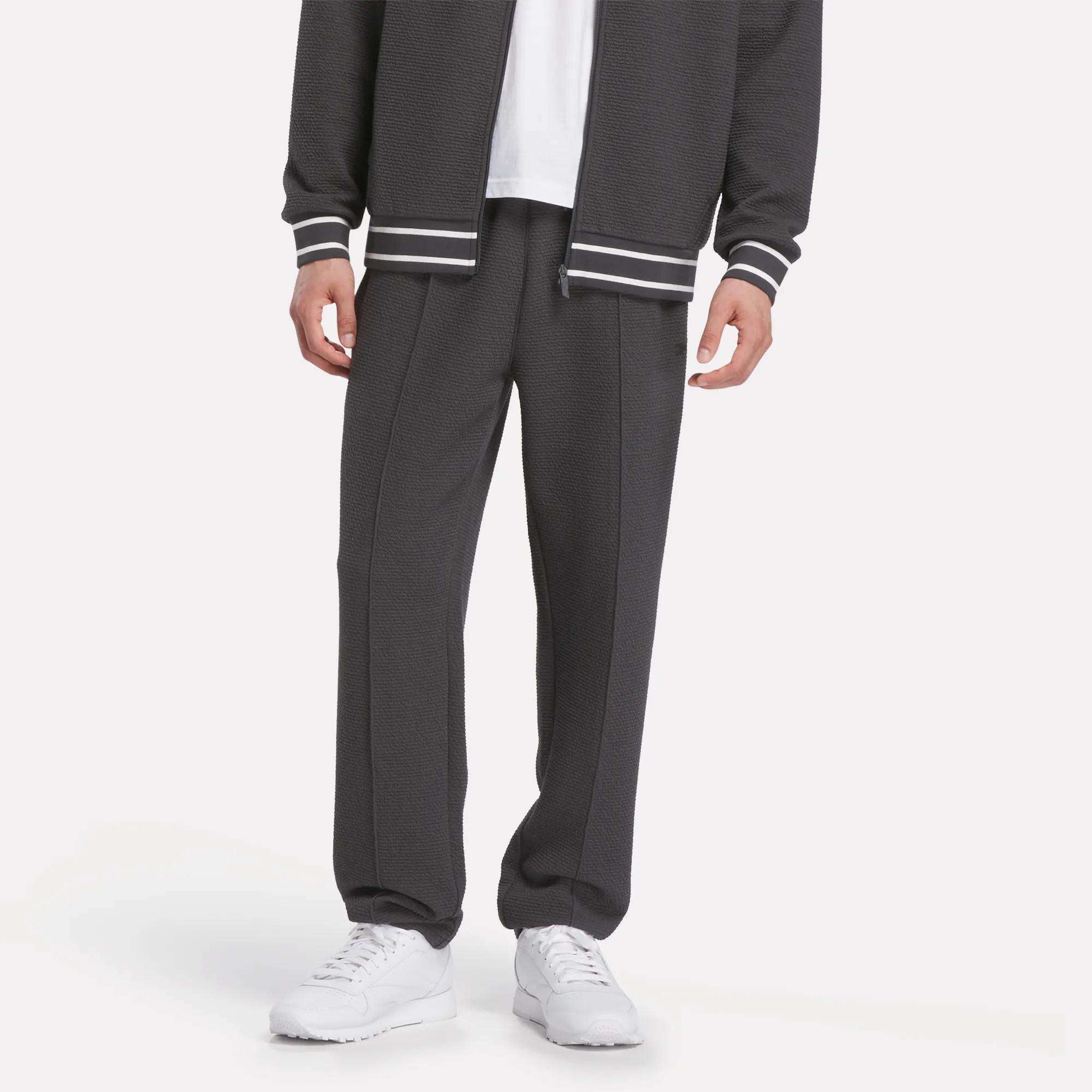 A man models Reebok's Men's Club Sport Textured Pants in dark gray with a slim tapered fit, paired with a matching jacket, white shirt, and white athletic shoes, shown shoulders down against a plain light background.