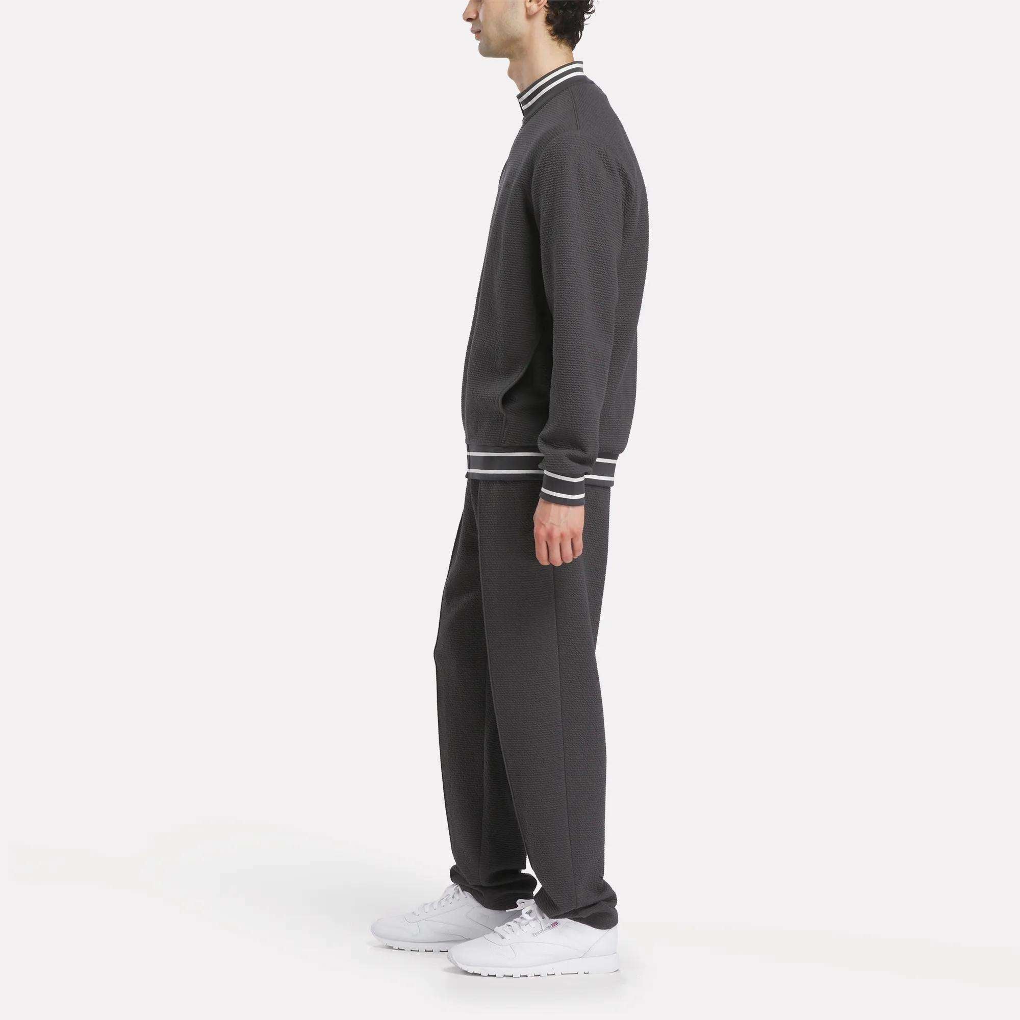 A man stands in profile wearing Reebok Men's Club Sport Textured Pants in dark gray with a matching top featuring white and black striped cuffs and collar, paired with white sneakers, against a plain white background.
