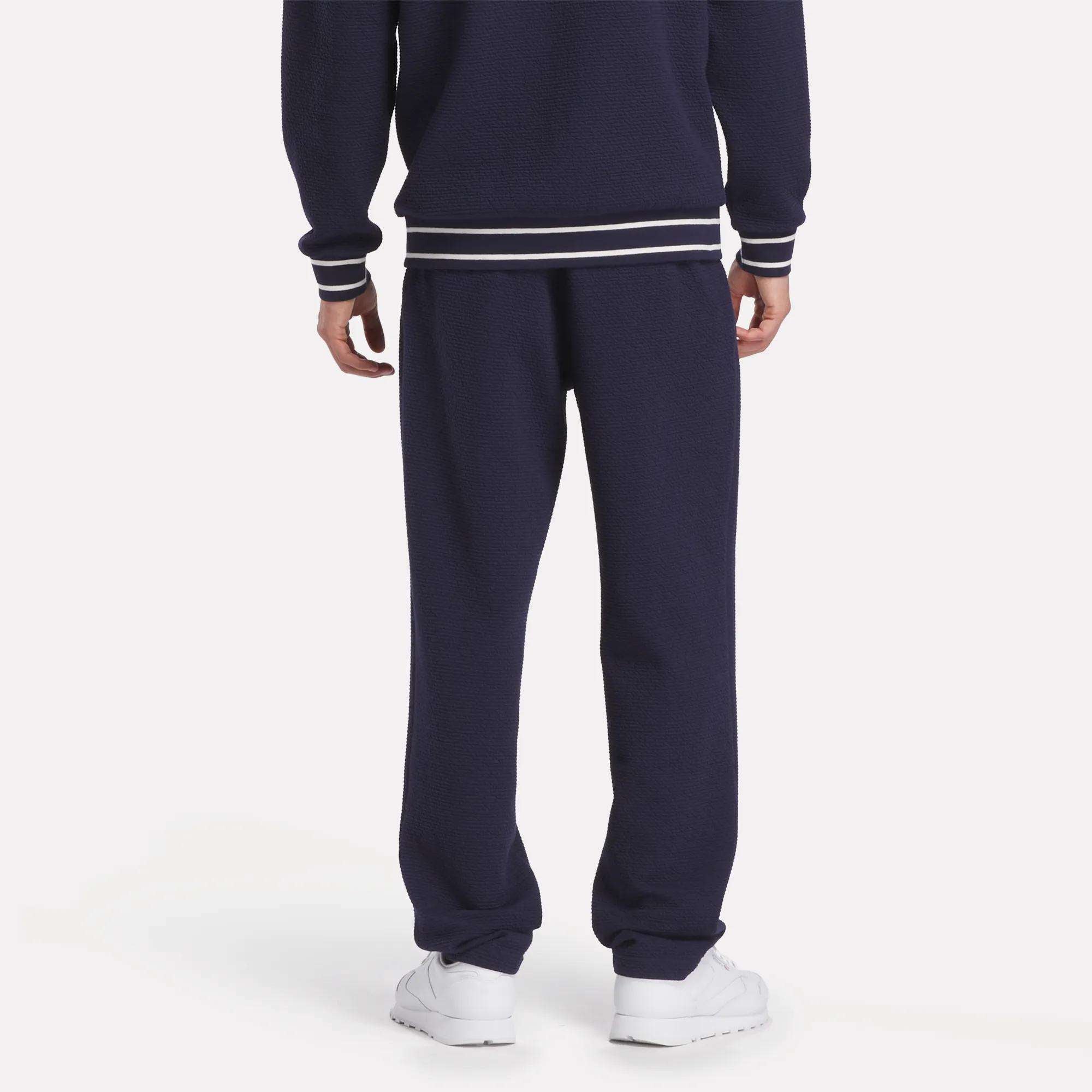 A person faces away, wearing Reebok Men's Club Sport Textured Pants in navy blue with white-striped cuffs and hem, paired with white sneakers—ideal for cool weather—set against a plain white background.