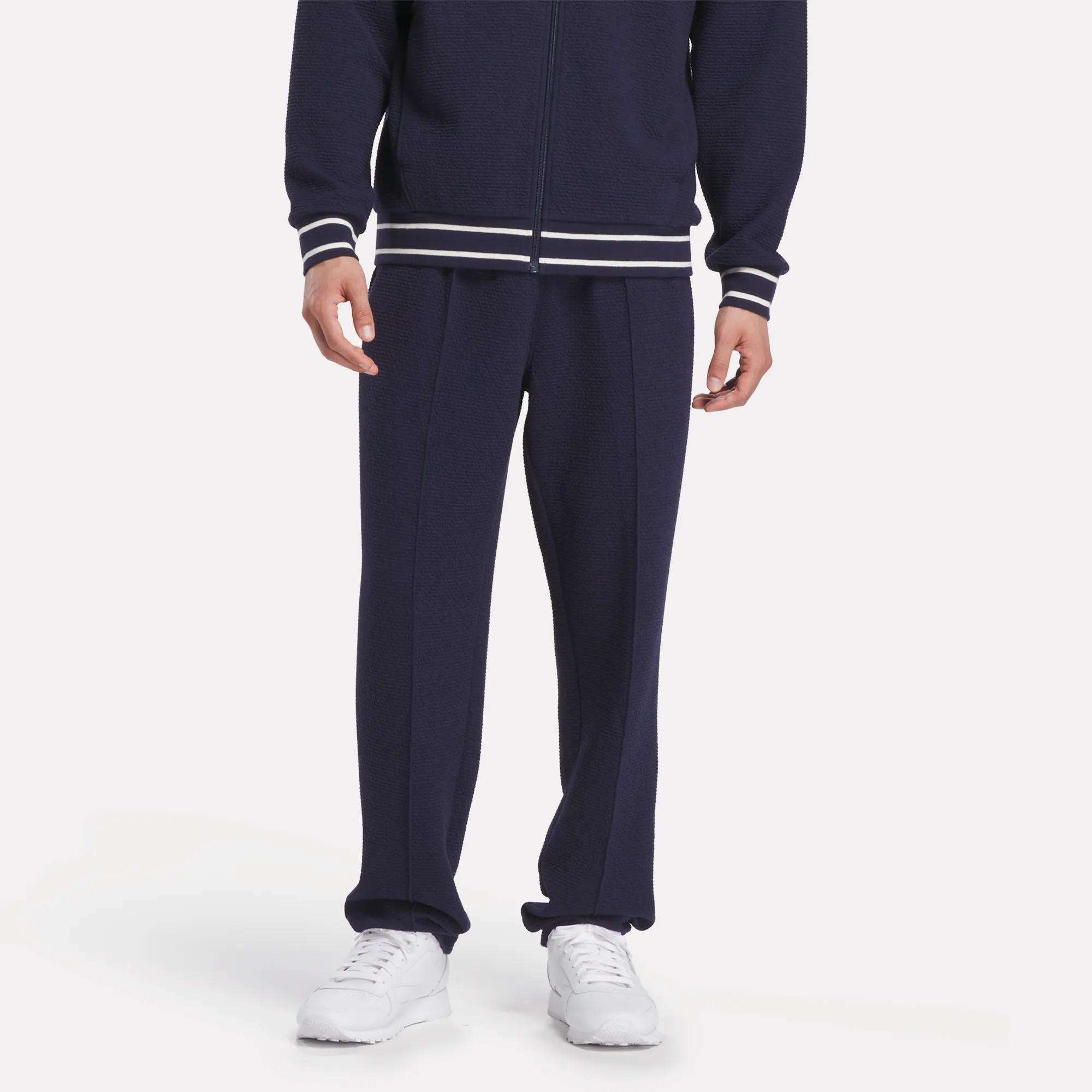 A person wearing Reebok Men's Club Sport Textured Pants—navy blue track pants with a matching zip-up jacket, white striped cuffs, and white sneakers—stands against a plain background. Their face is not visible.