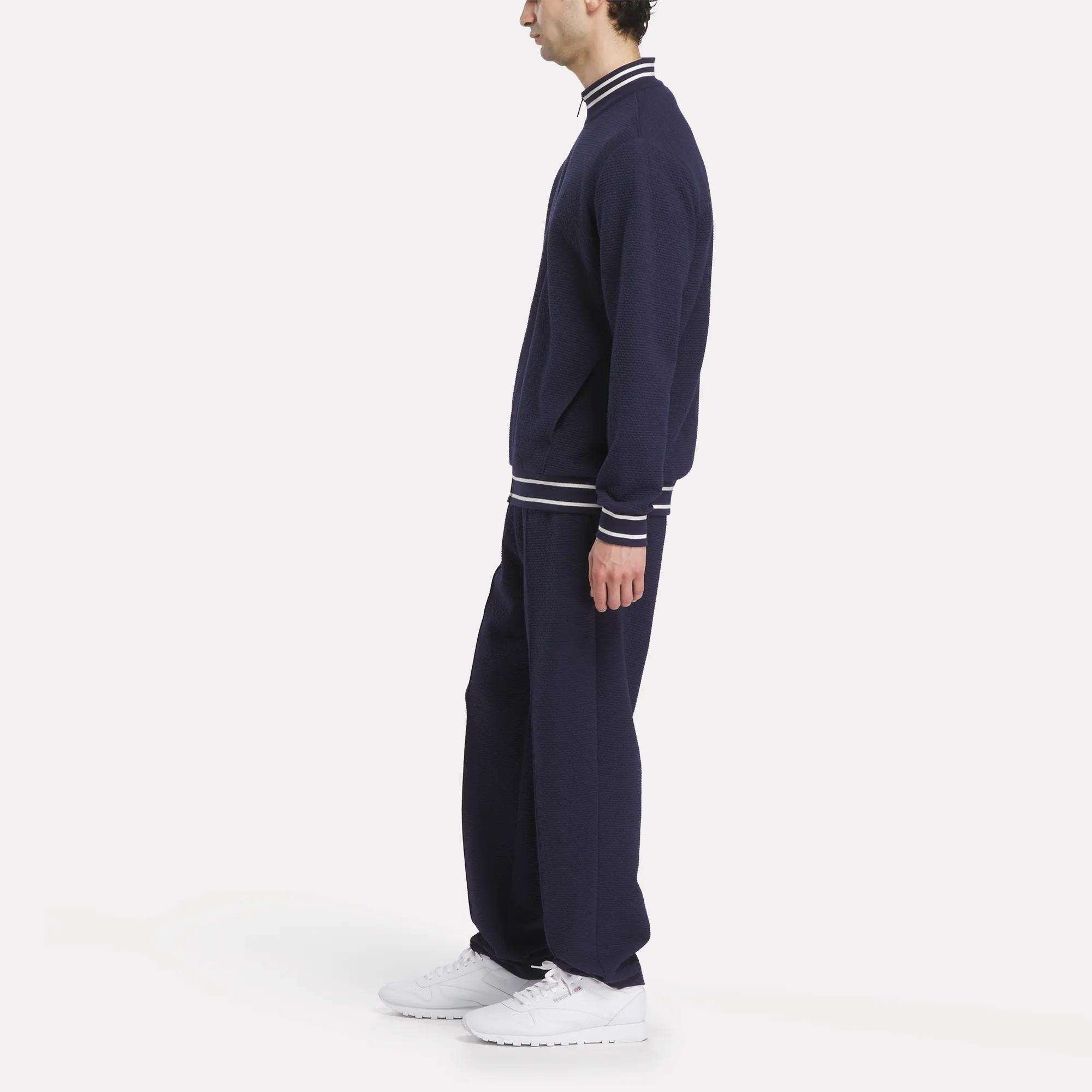 A man stands in profile against a white background, wearing Reebok Men's Club Sport Textured Pants as part of a navy blue tracksuit with white trim on the cuffs and collar, paired with white sneakers.
