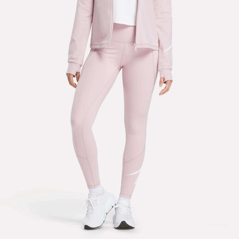 A person stands against a plain white background wearing Reebok Women's High-Rise Tights 26", a light pink athletic jacket, and white sneakers. The individual's head is not visible in the image.