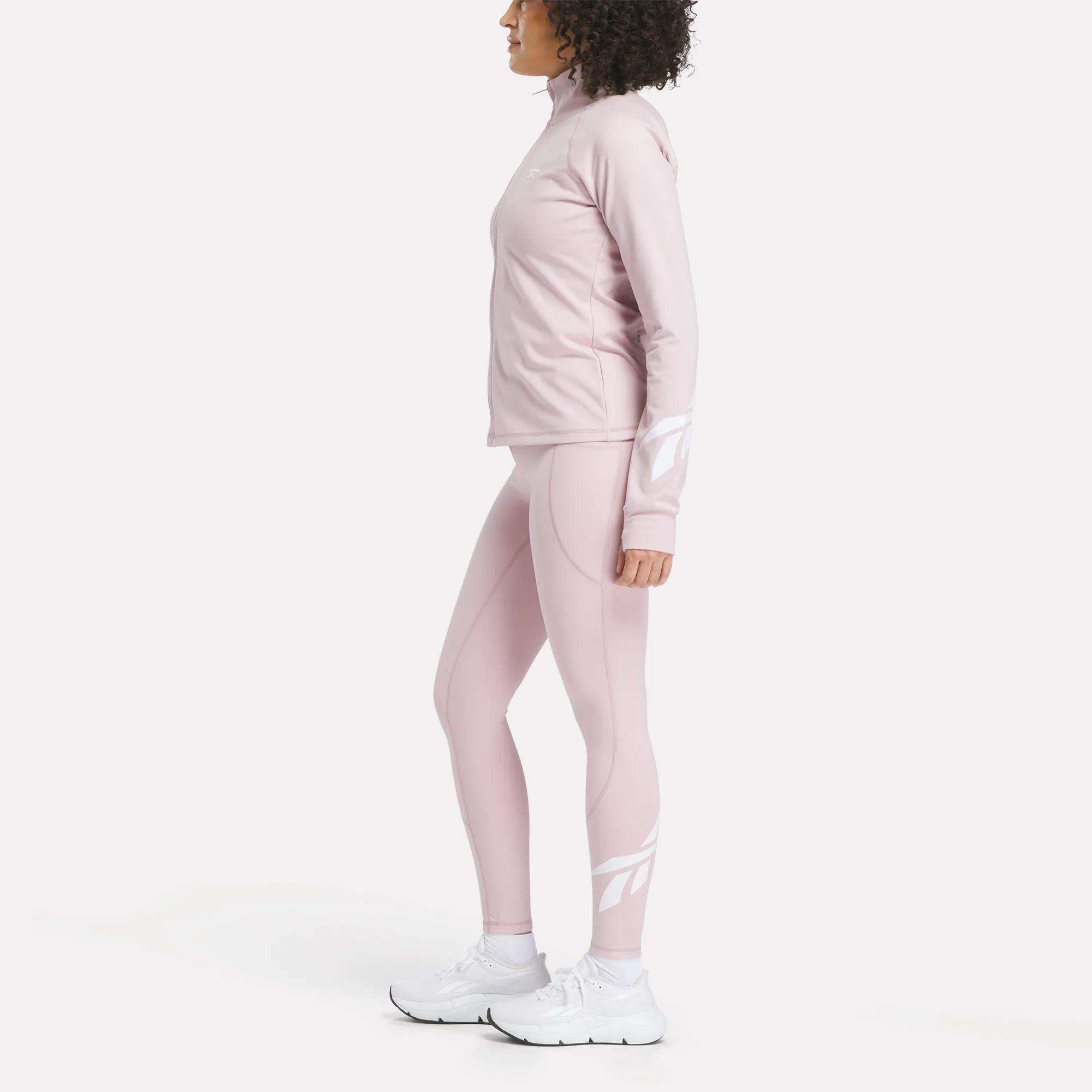 A woman stands in profile against a plain white background, wearing Reebok Women's High-Rise Tights 26" in light pink with long sleeves and white sneakers.