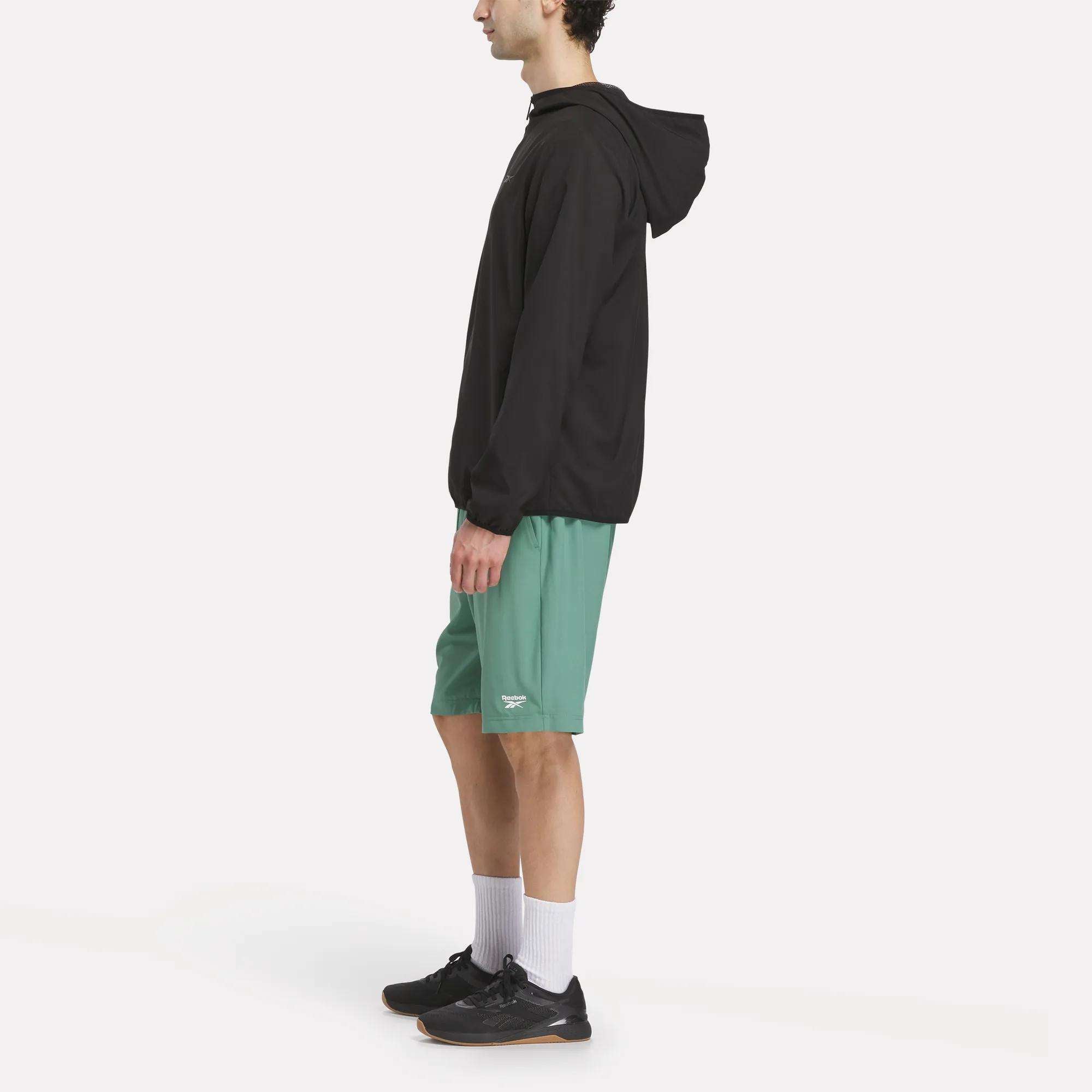 A person standing sideways in a Reebok Men's Woven Jacket, green shorts, white crew socks, and black athletic shoes against a plain white background.