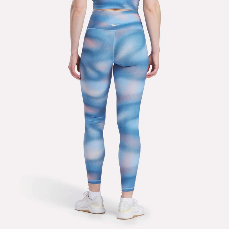 A woman faces away, wearing Reebok Women's Fluid Print High-Rise Tights with a matching top and white sneakers, all set against a plain white background.