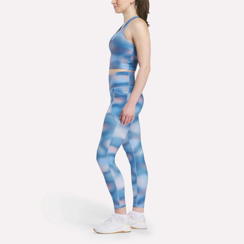 A woman in profile wears a Reebok Women's Cropped Bra Tank Top and matching blue-pink patterned leggings with light-impact support, paired with white athletic shoes, all set against a plain white background.