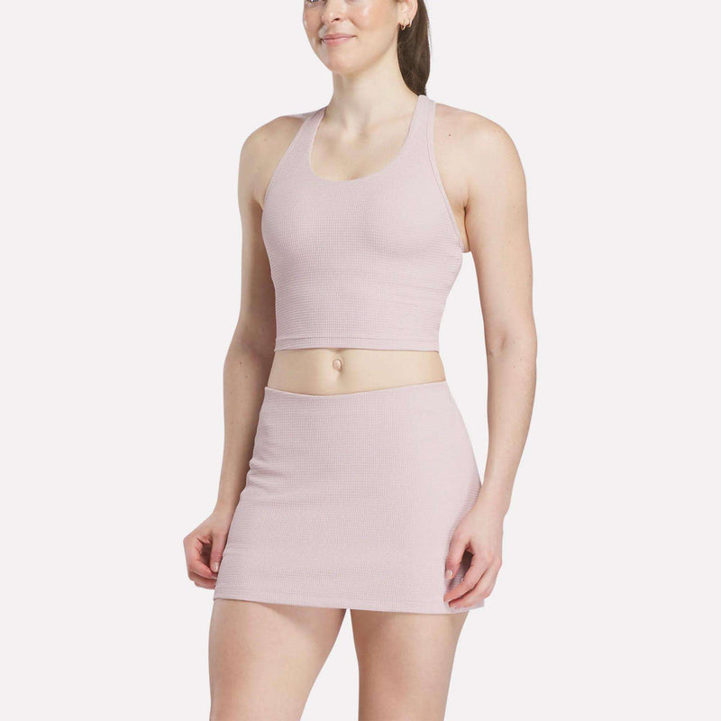 A woman in a light pink Women's Time Out Cropped Tank Top by Reebok and a matching high-waisted mini skirt stands against a plain white background, smiling slightly.