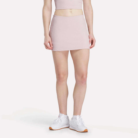 A person models the Reebok Women's Time Out Slim Tennis Skort in light pink with a matching crop top and white sneakers, standing against a plain white background. The individual's head is cropped from the image.