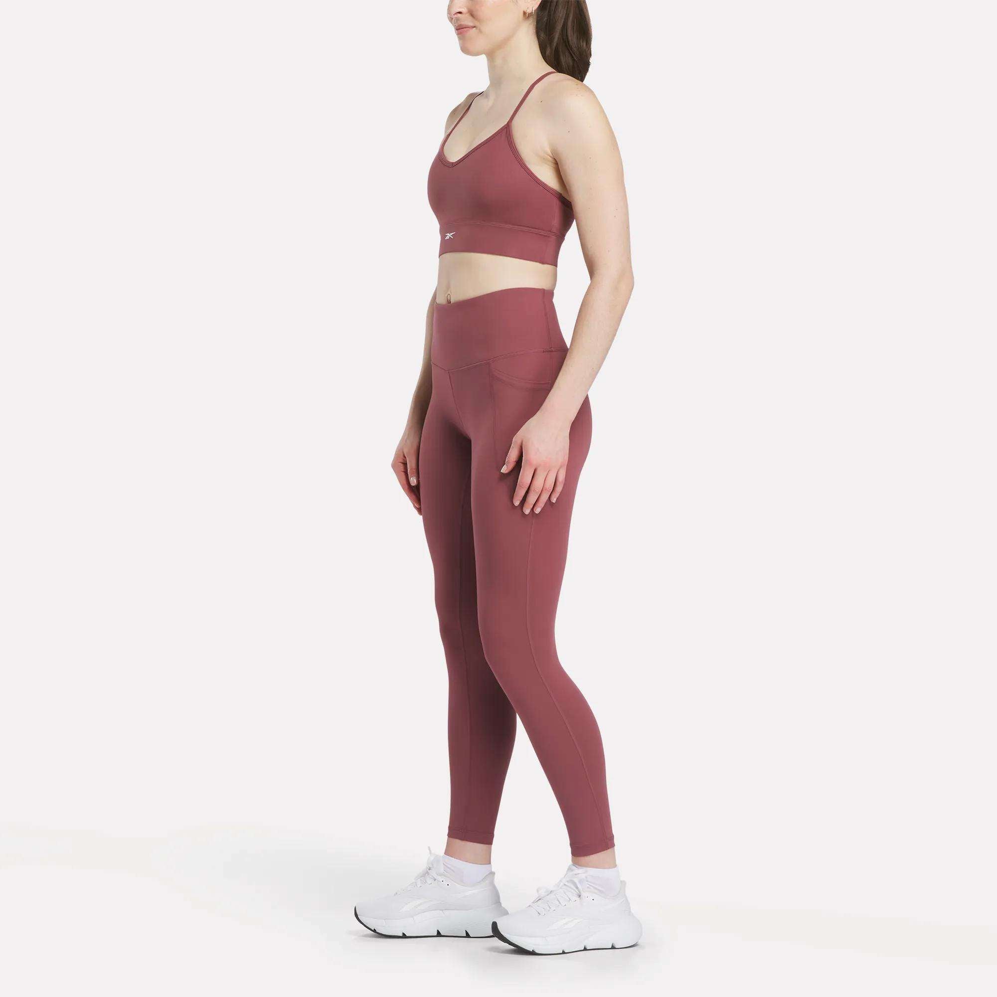 A woman in profile models Reebok Women's High-Rise Tights with a coordinating mauve sports bra and white athletic shoes, set against a plain white background.