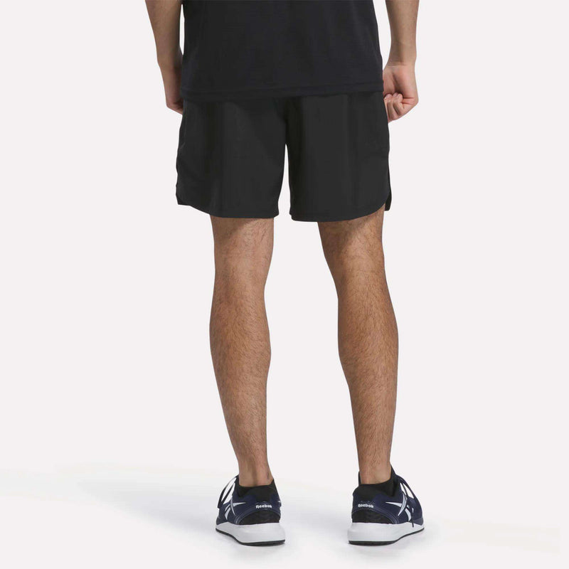 A person stands facing away, wearing a black shirt, Reebok Men’s Running Shorts 7″, and black sneakers with white soles, against a plain light background.