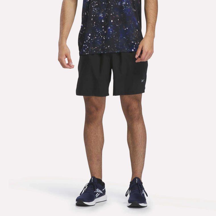 A person stands against a plain white background, wearing a black star-patterned shirt, black Reebok Men’s Running Shorts 7″, and navy blue running shoes. Only the lower half of their body is visible, highlighting the comfortable athletic fit of the Reebok shorts in black.
