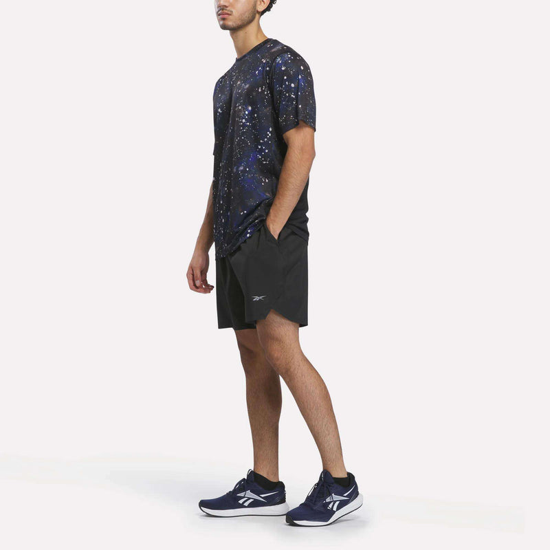 A man stands wearing a black galaxy-print t-shirt, Reebok Men’s Running Shorts 7″, and navy blue Reebok sneakers against a plain white background.