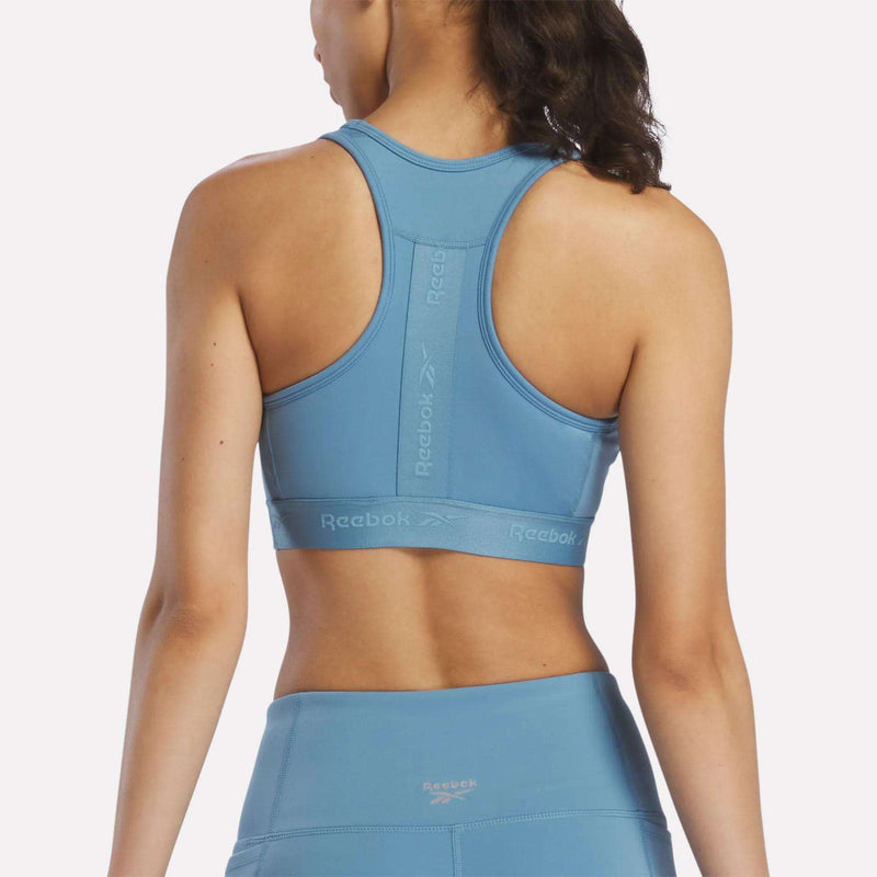 A woman is shown from the back wearing a light blue Reebok Women’s Medium-Impact Sports Bra with matching leggings. The sports bra features a racerback design, visible Reebok logo, and removable cups for customizable comfort.