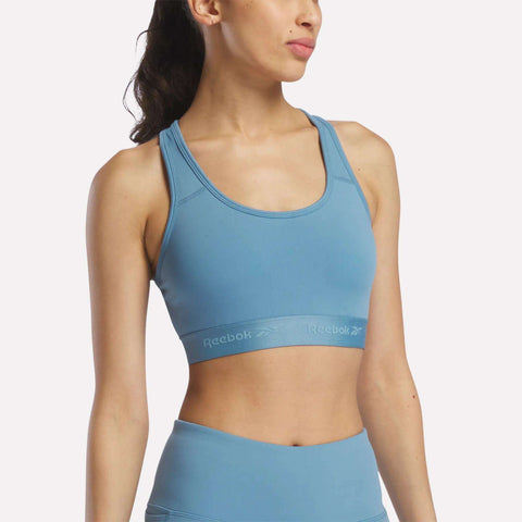 A woman stands against a plain background, facing slightly to the side with her head turned away from the camera, wearing a Reebok Women's Medium-Impact Sports Bra in Warped Blue with removable cups, paired with matching leggings.