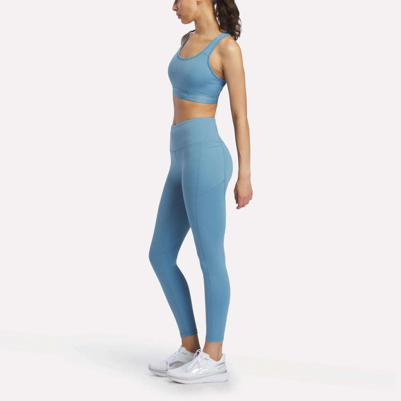 A woman is shown in profile against a plain white background, wearing a blue Reebok Women's Medium-Impact Sports Bra with matching leggings and white athletic shoes. The sports bra includes removable cups for customizable support.