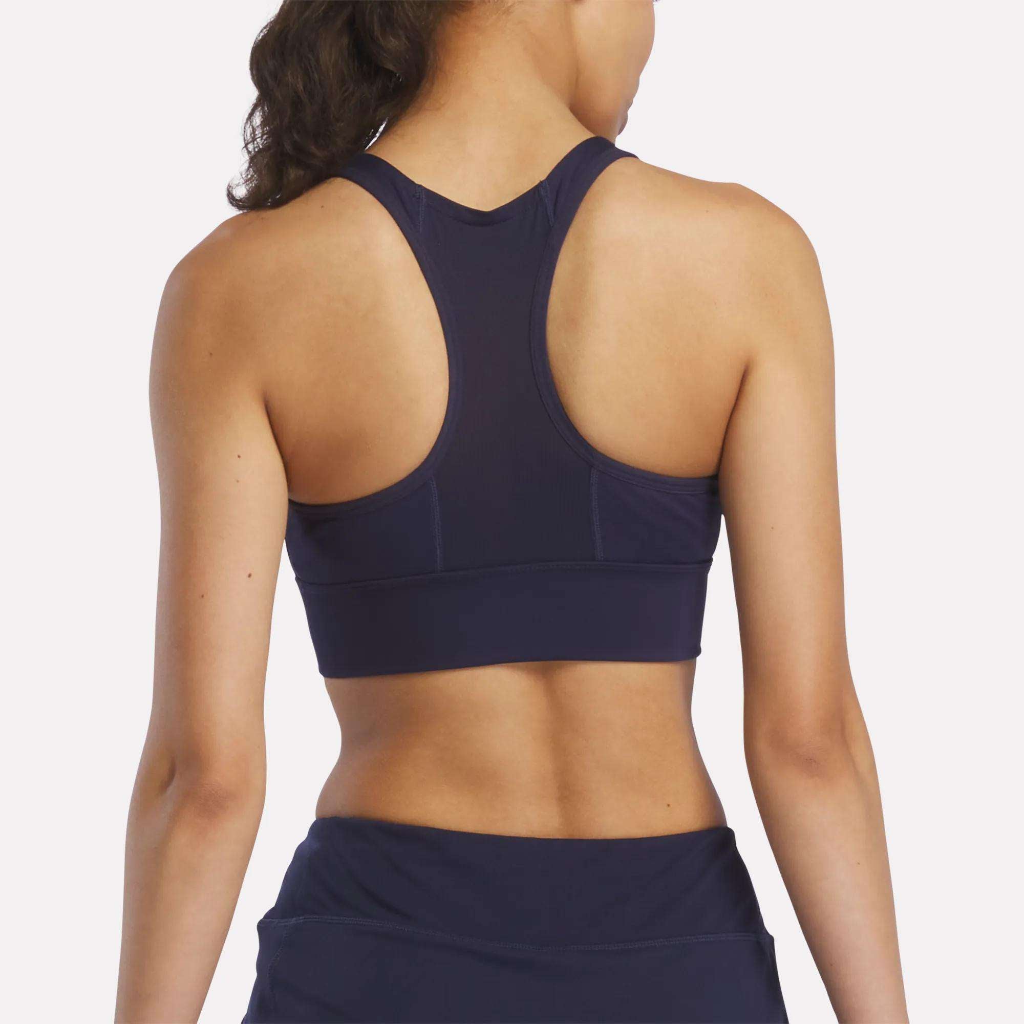 A woman with long dark hair in a ponytail is seen from behind wearing the Reebok Women's Rush Medium-Impact Sports Bra in navy blue with matching bottoms, perfect for medium-impact workouts, set against a plain light background.
