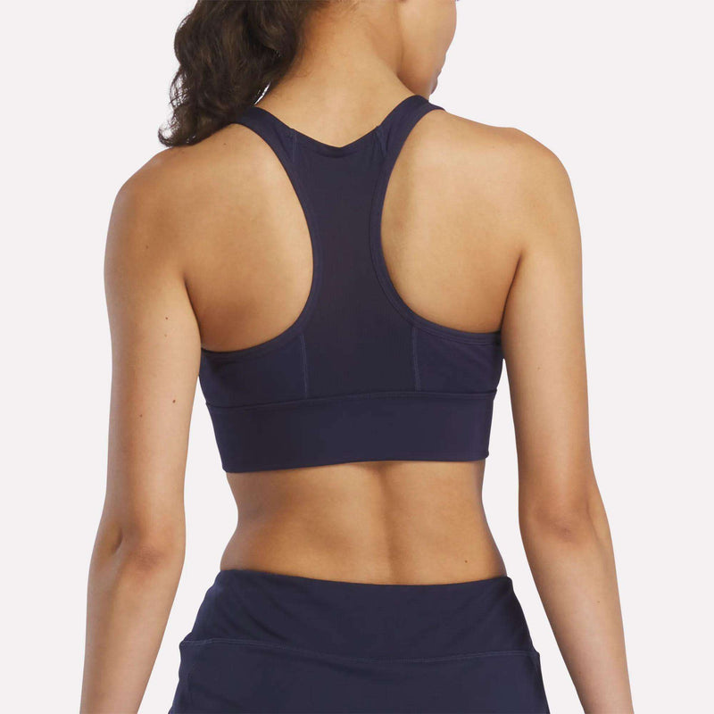 A woman with long dark hair in a ponytail is seen from behind wearing the Reebok Women's Rush Medium-Impact Sports Bra in navy blue with matching bottoms, perfect for medium-impact workouts, set against a plain light background.