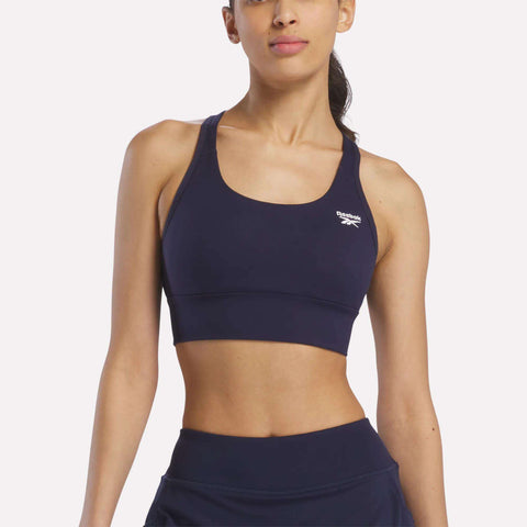 A woman stands against a plain light background wearing the Reebok Women's Rush Medium-Impact Sports Bra in navy blue with matching bottoms, ideal for medium-impact workouts.