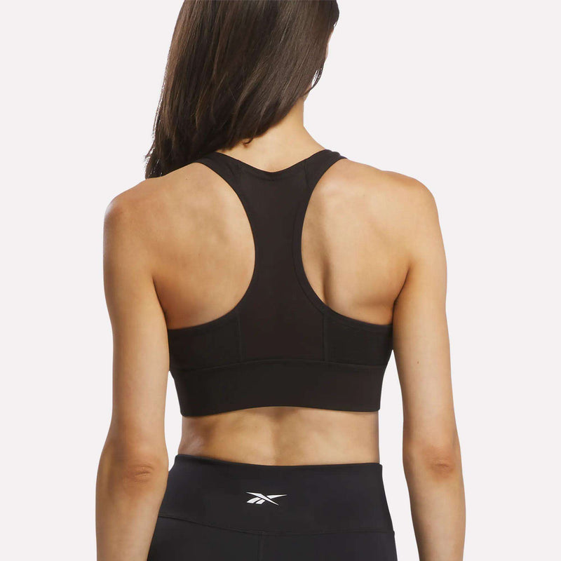 A woman with long brown hair is shown from the back against a plain white background wearing the Reebok Women's Rush Medium-Impact Sports Bra in black, paired with matching black leggings featuring the Reebok logo.
