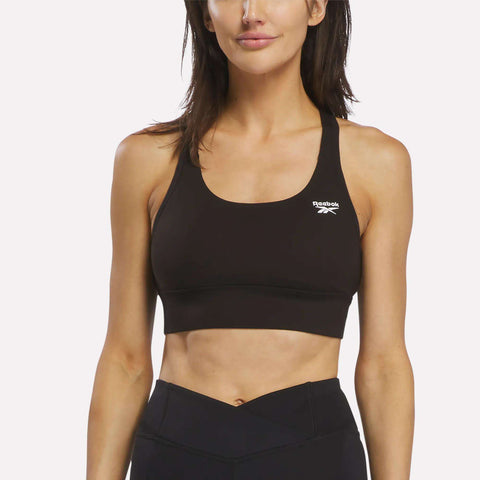 A woman stands facing forward against a plain white background, arms relaxed at her sides, wearing a black Reebok Women's Rush Medium-Impact Sports Bra and black workout leggings.