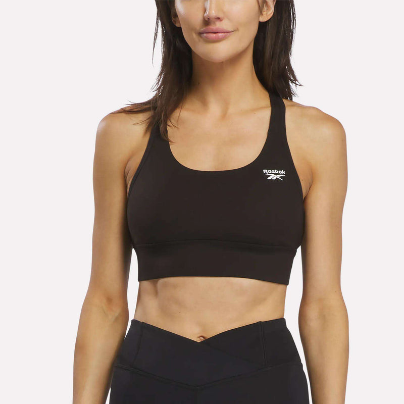 A woman stands facing forward against a plain white background, arms relaxed at her sides, wearing a black Reebok Women's Rush Medium-Impact Sports Bra and black workout leggings.