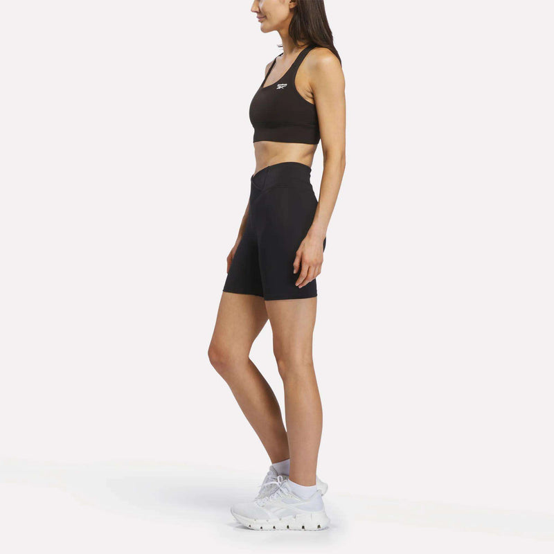 A woman stands in profile against a plain white background wearing the Reebok Women's Rush Medium-Impact Sports Bra in black, paired with black high-waisted biker shorts and white athletic shoes.