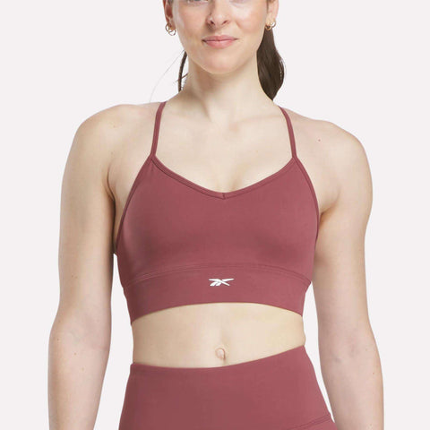 A woman models a mauve Reebok Women's Back Bra with thin straps and a small logo, paired with matching high-waisted leggings, standing against a plain white background.