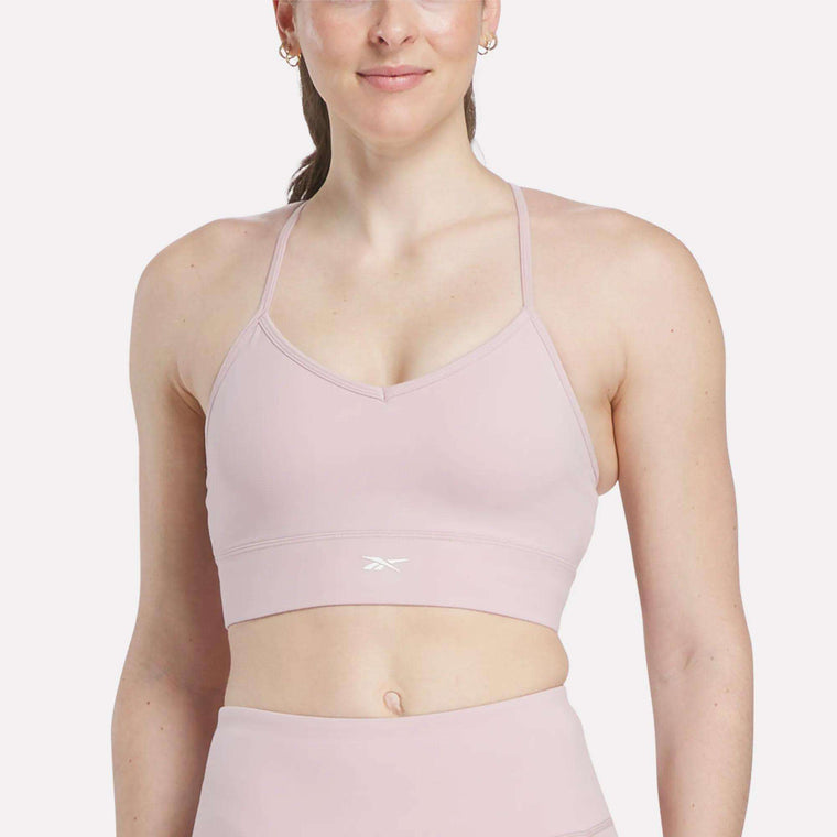 A woman wears the Reebok Women's Back Bra in light pink, paired with matching leggings, while standing against a plain white background. This sports bra provides medium-impact support for active comfort.