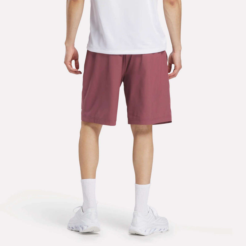 A person stands facing away from the camera against a plain background, wearing a white t-shirt, Reebok Men's Range Shorts 9" in mauve, white crew socks, and white sneakers.