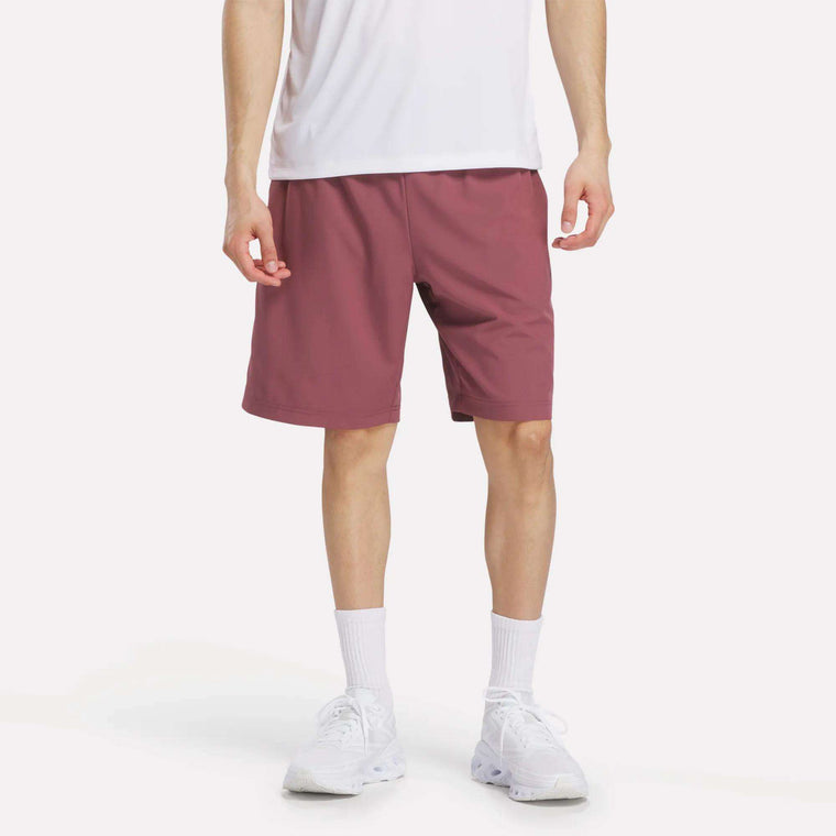 A person stands against a plain background wearing a white t-shirt, white crew socks, white sneakers, and mauve Reebok Men's Range Shorts 9". The photo is cropped at the shoulders.