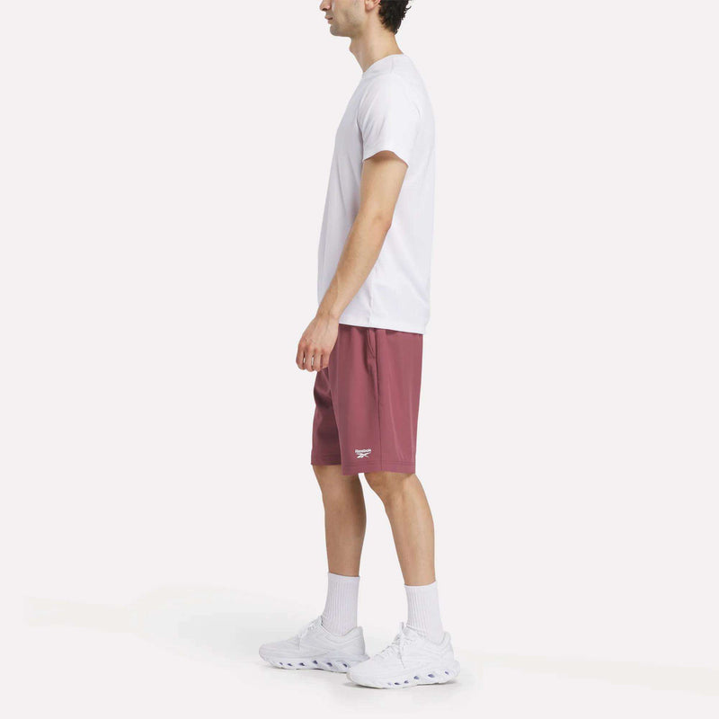 A person stands sideways against a white background, wearing a white T-shirt, Men's Range Shorts 9" by Reebok in maroon, white crew socks, and white sneakers.
