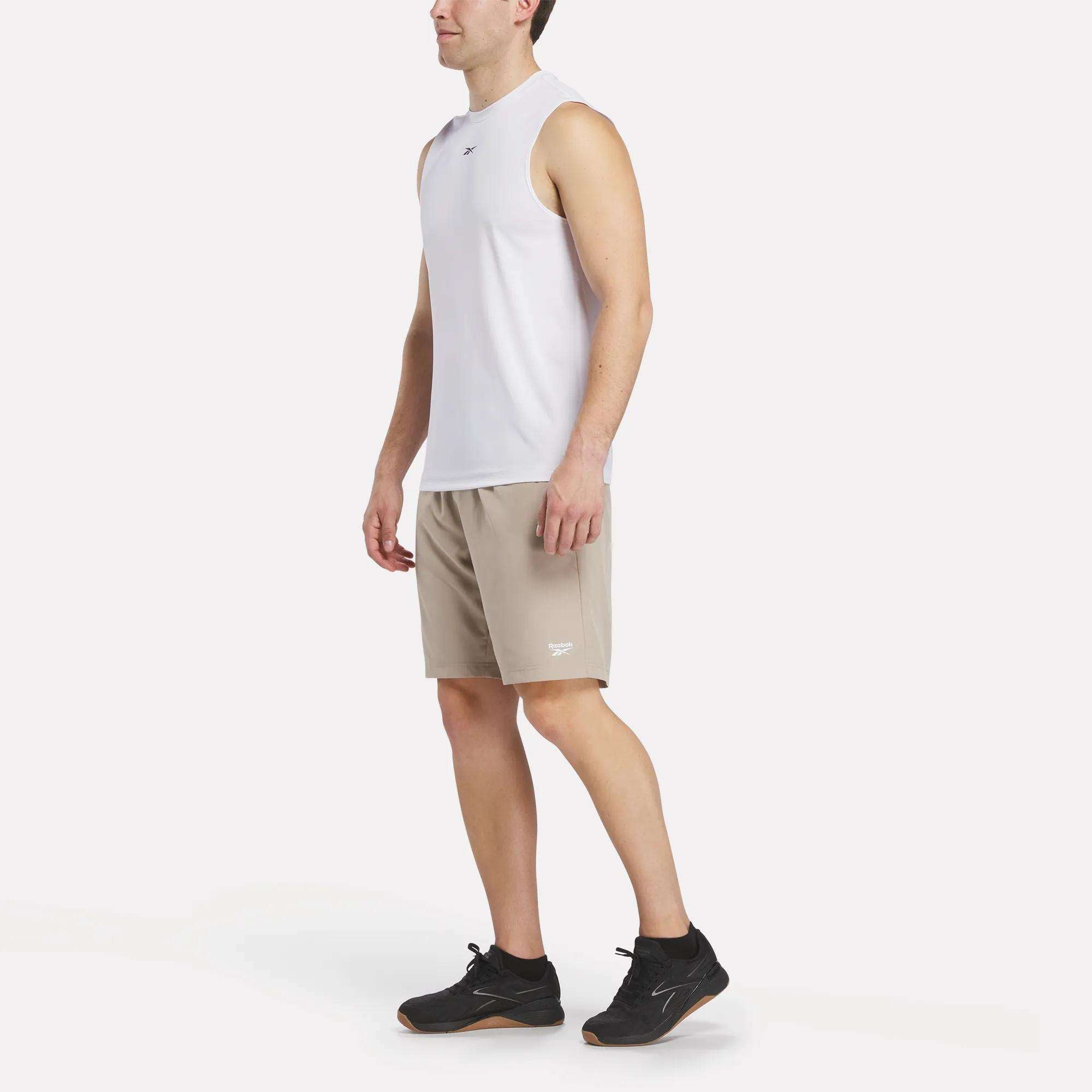 A man stands against a white background wearing a sleeveless white athletic shirt, black sneakers, and beige Reebok Men's Range Shorts 9", looking forward with his arms relaxed at his sides.