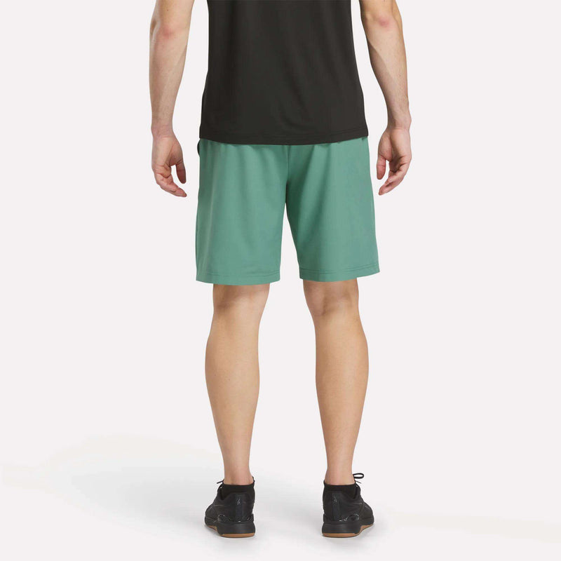 A person wearing a black shirt, Reebok Men’s Range Shorts 9″ in teal, and black shoes stands facing away from the camera against a white background.
