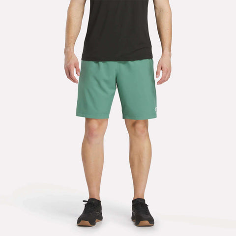 A person wearing a black shirt, Reebok Men’s Range Shorts 9″ in Field Green, and black athletic shoes stands against a plain, light background. Only the lower torso and legs are visible.