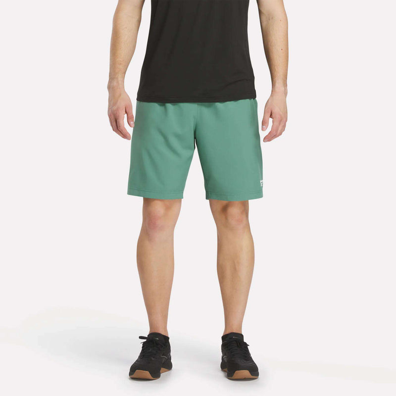 A person wearing a black shirt, Reebok Men’s Range Shorts 9″ in Field Green, and black athletic shoes stands against a plain, light background. Only the lower torso and legs are visible.