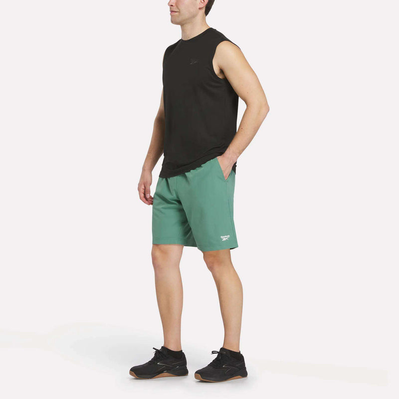 A person stands wearing a sleeveless black shirt, Reebok Men’s Range Shorts 9″ in green, and black sneakers against a plain white background.