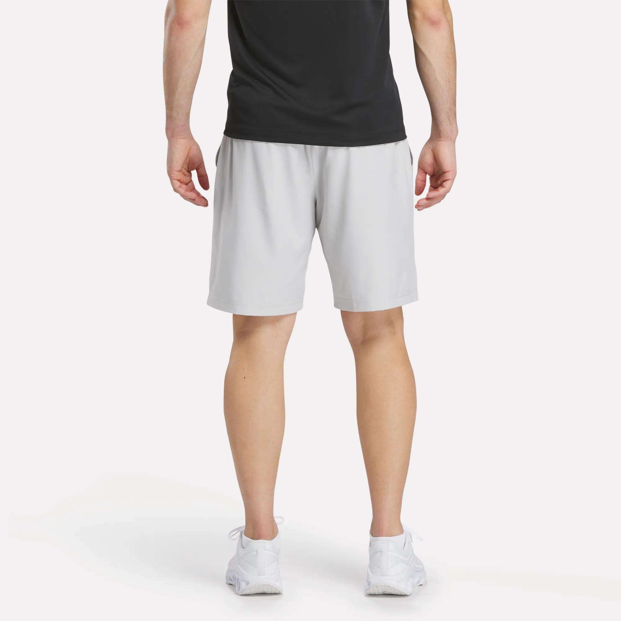 A person wearing a black shirt, white sneakers, and Reebok Men's Range Shorts 9" in light gray stands facing away from the camera against a plain white background.