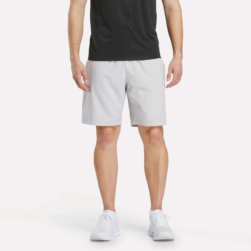 A man wearing a black T-shirt, Reebok Men’s Range Shorts 9″ in Grey 1, and white sneakers stands against a plain white background, facing forward. His head is not visible in the image.