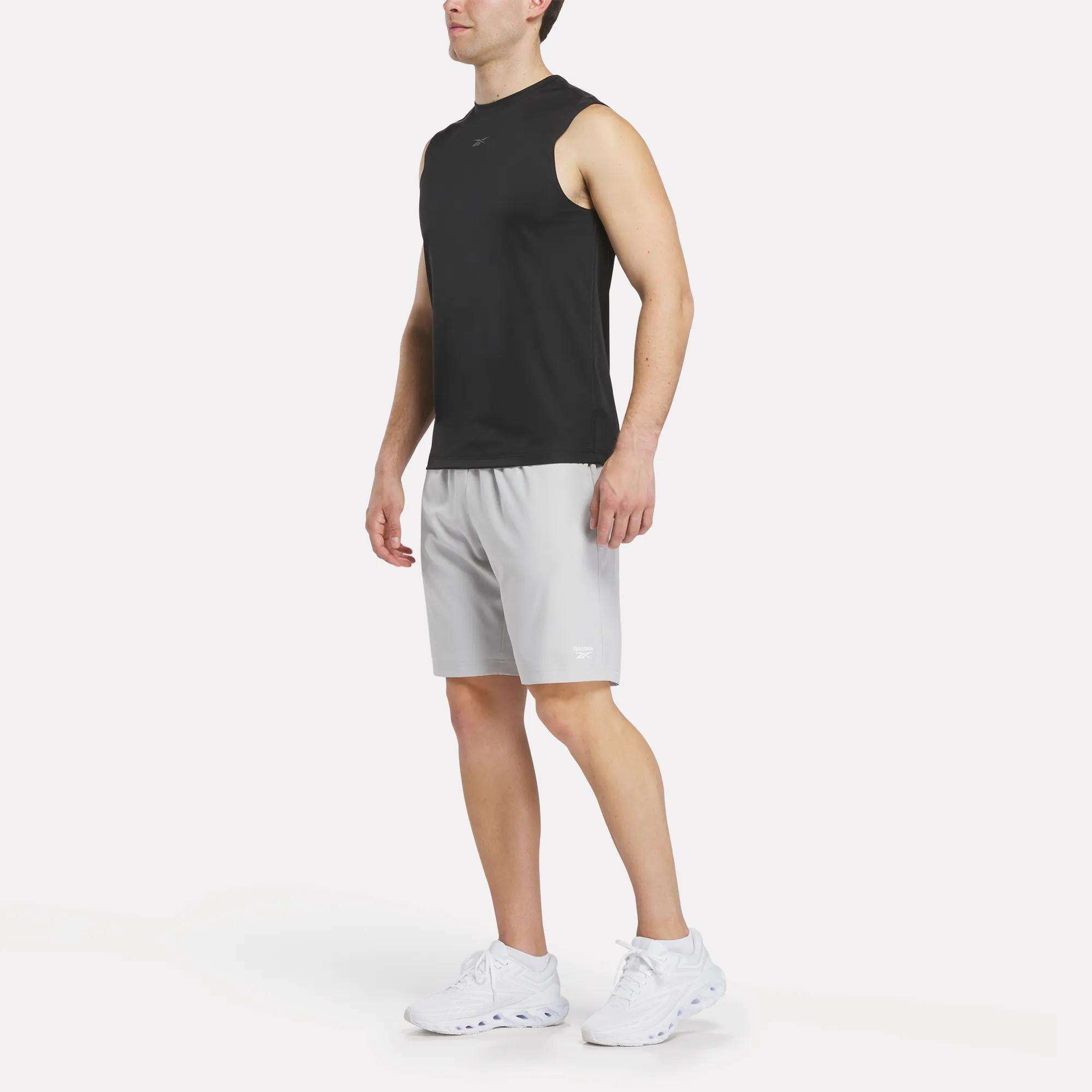 A man wearing a black sleeveless athletic shirt, Reebok Men's Range Shorts 9" in light gray, and white sneakers stands on a white background.