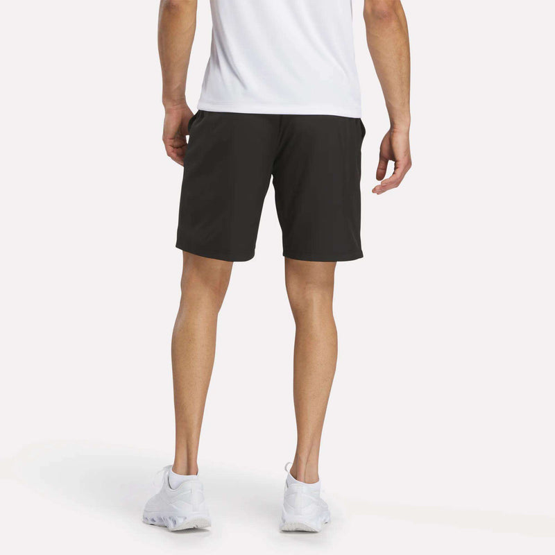 A person stands facing away, wearing a white shirt, Reebok Men’s Range Shorts 9″ in black, and white sneakers against a plain background.